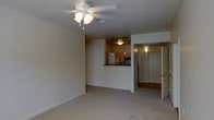 This single-floor residence offers a functional and cohesive layout with a calm, neutral aesthetic. The living area is finished with light wall-to-wall carpeting and painted in soft, warm tones, creating a welcoming atmosphere. A ceiling fan with a built-in light fixture serves as the central overhead lighting source, adding both practicality and a subtle design element to the space.

The kitchen is positioned in a semi-open configuration, separated from the living area by a raised dark-topped breakfast bar or counter partition. Inside, warm wood-toned cabinetry lines the walls, complemented by a set of pendant lights — including distinctive blue accent pendants — that hang from a track lighting system, adding visual interest. A full-sized refrigerator and additional appliances are visible within the kitchen, indicating a well-equipped cooking space.

A hallway visible from the living area leads to what appears to be a closet with mirrored sliding doors, suggesting access to a bedroom or storage area. The mirrored doors reflect light and contribute to a sense of added depth. Overall, the space maintains a clean, move-in-ready presentation with consistent finishes throughout.