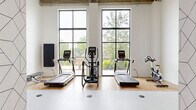 Located at 100 N Farwell St in Eau Claire, Wisconsin, this fitness center offers a well-appointed exercise space designed with both functionality and aesthetics in mind. The room is defined by its striking floor-to-ceiling windows framed in black steel, which flood the interior with abundant natural light and provide open views of the surrounding greenery and outdoor terrace. The exposed concrete ceiling adds an industrial character that contrasts beautifully with the crisp white walls and warm hardwood flooring found beneath the cardio equipment zone. A smooth, light-colored workout floor extends across the open central area, providing ample room for stretching, floor exercises, and functional training. Cardio equipment includes multiple treadmills, an elliptical trainer, and a stationary spin bike, all positioned to take advantage of the scenic window views. A dedicated free weights area features a fully stocked dumbbell rack alongside a wall-mounted mirror, enhancing the sense of space and supporting strength training needs. The entry area is distinguished by a geometric-patterned accent wall that adds a modern, design-forward touch to the space. A full-length mirror, weight bench, and balance equipment round out the fitness offerings. The overall ambiance is bright, clean, and motivating.
