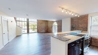 This two-story, two-bedroom, two-bathroom space offers a compelling mix of historic character and contemporary design across two well-proportioned floors. The main level centers around an open-plan kitchen and living area that immediately impresses with its exposed brick walls, expansive floor-to-ceiling windows, and rich hardwood flooring. The kitchen is well-appointed with dark espresso cabinetry, granite countertops, stainless steel appliances, and a breakfast bar peninsula that connects seamlessly to the living space. A dramatic arched window floods the living area with natural light and adds a striking architectural focal point. The lower level also includes a hallway with louvered closet doors and a full bathroom featuring a single vanity with dark wood cabinetry and tile flooring. Upstairs, the second floor houses both bedrooms, each finished with plush carpet and clean, neutral walls. The primary bedroom is generously sized and connects to a dedicated full bathroom complete with a double-sink vanity, dark wood cabinetry, a large mirror, and a tiled tub-shower combination. The second bedroom is comfortable and well-lit. Throughout the space, the interplay of exposed brick, modern fixtures, and warm wood tones creates a distinctive urban aesthetic with a welcoming atmosphere.