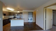 This single-floor space is designed with an open-concept layout that seamlessly connects the kitchen and living areas, creating a cohesive and functional environment. The kitchen is outfitted with stainless steel appliances, including a refrigerator, stove, and microwave, complemented by dark wood cabinetry and a light granite-topped island or peninsula that serves as both a workspace and a casual dining area. A pendant light hangs above, adding a focused point of illumination to the kitchen zone.

The flooring throughout the main living area consists of polished concrete, giving the space an industrial yet modern aesthetic that is both durable and easy to maintain. The neutral-toned walls and clean-lined architecture contribute to a minimalist ambiance throughout.

A notable feature is the in-unit laundry closet, visible through a sliding or open doorway, which houses a full-size front-loading washer and dryer along with shelving for storage. This dedicated utility space adds a high level of convenience. The overall design prioritizes practicality and modern style, making efficient use of the available space.