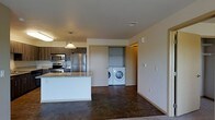 This single-floor space is designed with an open-concept layout that seamlessly connects the kitchen and living areas, creating a cohesive and functional environment. The kitchen is outfitted with stainless steel appliances, including a refrigerator, stove, and microwave, complemented by dark wood cabinetry and a light granite-topped island or peninsula that serves as both a workspace and a casual dining area. A pendant light hangs above, adding a focused point of illumination to the kitchen zone.

The flooring throughout the main living area consists of polished concrete, giving the space an industrial yet modern aesthetic that is both durable and easy to maintain. The neutral-toned walls and clean-lined architecture contribute to a minimalist ambiance throughout.

A notable feature is the in-unit laundry closet, visible through a sliding or open doorway, which houses a full-size front-loading washer and dryer along with shelving for storage. This dedicated utility space adds a high level of convenience. The overall design prioritizes practicality and modern style, making efficient use of the available space.