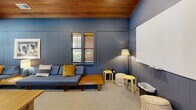 Located at 211 North Ave in Athens, Georgia, this thoughtfully designed living room offers a compelling blend of comfort and functionality. The space is immediately distinguished by its deep blue vertical shiplap walls, which lend a bold, cohesive character throughout. Overhead, a striking warm-toned wood-paneled vaulted ceiling adds architectural depth and a sense of elevated height, complemented by recessed lighting and a ceiling fan for year-round comfort.

Natural light filters in through multiple windows dressed with wooden blinds, offering views of the surrounding greenery. The room is furnished with low-profile blue mid-century modern sofas accented by rich golden-yellow throw pillows, creating a vibrant yet harmonious color palette. A large whiteboard mounted on one wall signals the room's versatility, making it equally suited for collaborative work, presentations, or casual gatherings.

Flexible seating options — including lightweight stools, poufs, and movable tables — allow the layout to adapt to a variety of uses. Soft carpeting underfoot adds warmth and comfort, while a ceiling fan and tasteful floor lamp round out the well-appointed interior. The space connects openly to adjacent areas, enhancing its sense of flow and accessibility.