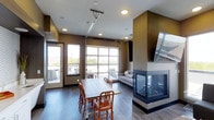 Located at 2900 East 16th Avenue in Denver, Colorado, this single-floor communal space combines contemporary design with functional comfort. The open-plan interior is anchored by a striking three-sided glass fireplace, serving as a central focal point that separates the dining and lounge areas while allowing visibility and warmth throughout. The dining area features a large farmhouse-style table with mixed wooden chairs, ideal for group gatherings. A full-panel glass garage door dominates one wall, flooding the interior with natural light and opening seamlessly to an outdoor patio area furnished with seating — perfect for indoor-outdoor entertaining. The lounge zone offers a comfortable sofa beneath a wall-mounted flat-screen television, creating a relaxed media area. Rich dark wood-style flooring runs throughout, complementing the deep earth-toned walls that lend the space a warm, sophisticated ambiance. Recessed ceiling lighting provides even illumination across the space. A built-in exhaust hood hints at kitchen or catering functionality nearby. Overall, the space strikes a balance between modern industrial design and inviting warmth, making it well-suited for social events, meetings, or community use.