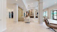 Located at 7430 Lyons Rd in Wellington, Florida, this sophisticated single-floor clubhouse spans a generously sized footprint designed for upscale communal living. The open-concept layout flows seamlessly from one lounge area to the next, united by large-format porcelain tile flooring and a bright, neutral palette throughout. Structural columns clad in warm wood-toned panels add architectural character while defining distinct seating zones without the use of dividing walls. 

The main lounge is a showpiece, anchored by a dramatic feature wall housing a large flat-screen display above a linear electric fireplace, flanked by built-in cabinetry with display niches. A separate sitting area showcases a glass-and-gold partition wall, creating a refined sense of enclosure with curated artwork and a marble-top coffee table. A moodier lounge nook features globe pendant lighting, bold art, and floor-to-ceiling windows offering lush views of palm trees and the pool beyond. 

The dining and bar area provides casual seating along a counter, while a separate dining space with natural wood tables opens directly to the outdoor pool area through expansive glass doors. Recessed lighting, statement chandeliers, and gold accents tie the design together, creating an upscale, resort-style ambiance throughout.