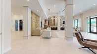 Located at 7430 Lyons Rd in Wellington, Florida, this sophisticated single-floor clubhouse spans a generously sized footprint designed for upscale communal living. The open-concept layout flows seamlessly from one lounge area to the next, united by large-format porcelain tile flooring and a bright, neutral palette throughout. Structural columns clad in warm wood-toned panels add architectural character while defining distinct seating zones without the use of dividing walls. 

The main lounge is a showpiece, anchored by a dramatic feature wall housing a large flat-screen display above a linear electric fireplace, flanked by built-in cabinetry with display niches. A separate sitting area showcases a glass-and-gold partition wall, creating a refined sense of enclosure with curated artwork and a marble-top coffee table. A moodier lounge nook features globe pendant lighting, bold art, and floor-to-ceiling windows offering lush views of palm trees and the pool beyond. 

The dining and bar area provides casual seating along a counter, while a separate dining space with natural wood tables opens directly to the outdoor pool area through expansive glass doors. Recessed lighting, statement chandeliers, and gold accents tie the design together, creating an upscale, resort-style ambiance throughout.