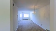 This single-floor space offers a clean, classic interior highlighted by a bright and open living area. The room is finished with soft, neutral-toned carpet that extends throughout, providing a warm and comfortable feel underfoot. Crisp white walls and white baseboards contribute to a light, airy atmosphere that feels fresh and well-maintained.

A standout feature of the living area is the elegant gold chandelier, which adds a traditional decorative touch and serves as a focal point for the room. Crown molding lines the ceiling, lending architectural character and a sense of refined detail to the otherwise simple, uncluttered space.

Natural light fills the room through a window positioned near the front entry door, which features a decorative glass insert that further enhances brightness. A baseboard heating system runs along the perimeter walls, a practical and efficient feature for year-round comfort. The entry area integrates seamlessly with the main living space, creating an open flow that makes the interior feel expansive.

Overall, the space presents a straightforward, move-in-ready layout with timeless design elements — ideal for those seeking a bright, neutral canvas that can be personalized to any style or furnishing preference.