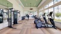 Located at 1575 League Line Rd in Conroe, Texas, this single-floor fitness center is designed to deliver a bright, motivating workout environment. The space is anchored by a distinctive tray ceiling accented in a soft sage green, complemented by a ceiling fan and recessed lighting that enhance the room's airy atmosphere. Patterned, wood-tone carpet tiles cover the floor throughout, lending warmth and a clean aesthetic to the space.

Large windows line multiple walls, flooding the interior with natural light and offering views of the surrounding outdoor pool area and towering pine trees — a defining feature of the East Texas landscape. Full-length mirrors along one wall visually expand the space and support proper form during workouts.

The fitness center is comprehensively equipped with a wide array of cardio machines, including treadmills, ellipticals, and stationary cycling bikes positioned to take advantage of the outdoor views. Strength training is well-represented with a multi-functional cable machine, a weight rack with barbells and plates, and a tiered dumbbell rack. A mounted flat-screen television provides entertainment during workouts. Glass-paneled doors offer easy entry and exit, contributing to the open, connected feel of this well-appointed fitness facility.
