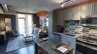 This single-floor space makes a strong visual impression through its thoughtful design and bold use of color. A striking burnt-orange accent wall anchors the open-concept layout, adding warmth and energy throughout. The kitchen is outfitted with sleek, contemporary grey cabinetry that extends floor to ceiling, maximizing storage while maintaining a clean, modern aesthetic. A geometric mosaic tile backsplash in grey and black tones adds texture and visual interest behind the sink and countertops. The kitchen flows naturally into a combined dining and living area, where a grey-toned dining table with place settings serves as a stylish transition between spaces. Dark hardwood flooring runs throughout, grounding the design with richness and continuity. The living area features a comfortable sofa, a low-profile coffee table, and carefully selected accent pieces that contribute to the space's urban, contemporary feel. Track lighting on the ceiling provides directional illumination, complementing the natural light that enters through large windows and a sliding glass door leading to a private balcony. The exposed concrete ceiling adds an industrial character that balances the warmer design elements, resulting in a space that feels both sophisticated and inviting.