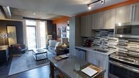 This single-floor space makes a strong visual impression through its thoughtful design and bold use of color. A striking burnt-orange accent wall anchors the open-concept layout, adding warmth and energy throughout. The kitchen is outfitted with sleek, contemporary grey cabinetry that extends floor to ceiling, maximizing storage while maintaining a clean, modern aesthetic. A geometric mosaic tile backsplash in grey and black tones adds texture and visual interest behind the sink and countertops. The kitchen flows naturally into a combined dining and living area, where a grey-toned dining table with place settings serves as a stylish transition between spaces. Dark hardwood flooring runs throughout, grounding the design with richness and continuity. The living area features a comfortable sofa, a low-profile coffee table, and carefully selected accent pieces that contribute to the space's urban, contemporary feel. Track lighting on the ceiling provides directional illumination, complementing the natural light that enters through large windows and a sliding glass door leading to a private balcony. The exposed concrete ceiling adds an industrial character that balances the warmer design elements, resulting in a space that feels both sophisticated and inviting.