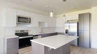 This well-designed 1-bedroom, 1-bathroom unit offers a thoughtful, efficient layout across a single floor. The open-concept kitchen serves as the heart of the space, showcasing a two-tone design with crisp white upper cabinetry and dark lower cabinets. A central island with a white quartz countertop provides additional workspace and casual seating. Stainless steel appliances, including a French door refrigerator with water dispenser, electric range, over-the-range microwave, and dishwasher, complete the contemporary kitchen. Cylindrical pendant lights hang above the island, adding a refined finishing touch. Light-toned hardwood flooring flows throughout the main living area, complementing the neutral, bright color palette. A sliding door offers access to an outdoor area and allows natural light to fill the space. The bedroom features soft carpeting, a window with outdoor views, and clean, neutral walls that create a calm and restful environment. A dedicated closet provides convenient storage. The full bathroom is generously proportioned, featuring a soaking tub, a separate walk-in shower, a floating dark vanity with white countertop and undermount sink, and light hardwood-style flooring, delivering a clean and functional design throughout.