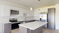 This well-designed 1-bedroom, 1-bathroom unit offers a thoughtful, efficient layout across a single floor. The open-concept kitchen serves as the heart of the space, showcasing a two-tone design with crisp white upper cabinetry and dark lower cabinets. A central island with a white quartz countertop provides additional workspace and casual seating. Stainless steel appliances, including a French door refrigerator with water dispenser, electric range, over-the-range microwave, and dishwasher, complete the contemporary kitchen. Cylindrical pendant lights hang above the island, adding a refined finishing touch. Light-toned hardwood flooring flows throughout the main living area, complementing the neutral, bright color palette. A sliding door offers access to an outdoor area and allows natural light to fill the space. The bedroom features soft carpeting, a window with outdoor views, and clean, neutral walls that create a calm and restful environment. A dedicated closet provides convenient storage. The full bathroom is generously proportioned, featuring a soaking tub, a separate walk-in shower, a floating dark vanity with white countertop and undermount sink, and light hardwood-style flooring, delivering a clean and functional design throughout.