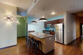 This well-appointed single-floor space offers a thoughtfully designed layout encompassing a combined kitchen and living room, a hallway, a full bathroom, and a closet. The open-concept kitchen and living area serves as the heart of the space, featuring rich dark hardwood flooring that flows throughout. The kitchen is equipped with stainless steel appliances — including a refrigerator, range, and microwave — complemented by dark wood cabinetry, granite countertops, and a large central island with seating for multiple guests. Striking dome pendant lights hang above the island, adding a modern industrial touch. Bold accent walls in shades of teal, turquoise, and sage green lend the space a vibrant, contemporary character. The living area is highlighted by wall-mounted sconce lighting and provides direct access to an outdoor area through large sliding glass doors, flooding the room with natural light. The hallway connects the main living areas to the bathroom, which features tile flooring, a vanity with dark wood cabinetry, and a shower-tub combination. The overall ambiance is modern and energetic, combining vivid color choices with sleek finishes and practical design to create a dynamic and functional environment.