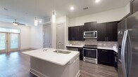 This single-floor unit showcases a contemporary design with a cohesive color palette of crisp whites, dark espresso cabinetry, and warm wood-tone flooring throughout. The open-concept kitchen serves as a central highlight, featuring a large white-topped center island with an integrated sink and pendant lighting above. Dark shaker-style cabinetry lines the walls, complemented by a stone tile backsplash and a full suite of stainless steel appliances, including a range, built-in microwave, and refrigerator. The kitchen flows naturally into the living area, which is finished with rich hardwood-style flooring, recessed lighting, and a ceiling fan. French doors with transom windows invite natural light and provide access to an outdoor space. The bedrooms are carpeted for comfort, each featuring ceiling fans and windows with blinds that offer natural light. The bathroom presents a clean, neutral aesthetic with large-format tile flooring, a tiled combination tub and shower, a dark-framed vanity mirror, and a sleek single-basin vanity. Throughout the unit, the consistent use of modern finishes and neutral tones creates a polished and inviting atmosphere.