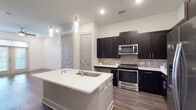 This single-floor unit showcases a contemporary design with a cohesive color palette of crisp whites, dark espresso cabinetry, and warm wood-tone flooring throughout. The open-concept kitchen serves as a central highlight, featuring a large white-topped center island with an integrated sink and pendant lighting above. Dark shaker-style cabinetry lines the walls, complemented by a stone tile backsplash and a full suite of stainless steel appliances, including a range, built-in microwave, and refrigerator. The kitchen flows naturally into the living area, which is finished with rich hardwood-style flooring, recessed lighting, and a ceiling fan. French doors with transom windows invite natural light and provide access to an outdoor space. The bedrooms are carpeted for comfort, each featuring ceiling fans and windows with blinds that offer natural light. The bathroom presents a clean, neutral aesthetic with large-format tile flooring, a tiled combination tub and shower, a dark-framed vanity mirror, and a sleek single-basin vanity. Throughout the unit, the consistent use of modern finishes and neutral tones creates a polished and inviting atmosphere.
