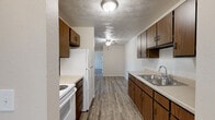 This single-floor unit is thoughtfully laid out across several rooms, each with distinct flooring and functional features. The galley-style kitchen runs along two parallel walls, lined with warm wood-toned cabinetry providing ample upper and lower storage. White appliances, including an electric range and refrigerator, are complemented by a stainless steel sink and light countertops. Wood-look vinyl plank flooring runs throughout the kitchen, which opens via a passthrough corridor into the main living areas. A ceiling fan is centrally mounted above.

The living areas are carpeted in a neutral tone and feature baseboard heating units along the walls. The main living room includes a full sliding glass door dressed with vertical blinds, allowing abundant natural light and outdoor views. A wall-mounted window AC unit provides additional climate control. The bedrooms are similarly carpeted with textured ceilings and individual windows fitted with horizontal blinds, each offering views of the surrounding greenery. The bathroom includes a full-size soaking tub with an integrated shower surround and chrome fixtures. Throughout the unit, textured ceilings and neutral wall tones contribute to a clean, functional aesthetic.