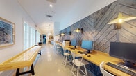 Located at 99 Bass Rd in Warner Robins, Georgia, this single-floor community clubhouse delivers a well-appointed, thoughtfully designed gathering space with distinct functional zones unified by a cohesive modern aesthetic. The grand entrance lobby immediately impresses with soaring vaulted ceilings, teal board-and-batten accent walls, and oversized pendant lights with gold interiors. Floor-to-ceiling glass doors flood the space with natural light while providing direct views of the outdoor pool area, and a striking chevron-patterned wood reception desk anchors the welcoming front area.

The adjoining lounge features large grid-pane windows that frame outdoor pool views, creating a bright and airy atmosphere. Comfortable seating, including a velvet sofa with mustard accent pillows, mid-century modern chairs, and curated artwork, gives this area a refined, residential feel.

A dedicated business center runs along one wall, lined with a continuous butcher-block countertop workstation set against a dramatic chevron-patterned reclaimed wood accent wall. Adjustable wall-mounted industrial sconces illuminate individual computer stations, while white bar-height chairs provide comfortable seating. A shuffleboard table adds a recreational element to this corridor.

A separate media lounge features a teal grid-paneled accent wall with mounted flat-screen televisions, stylish pendant lighting, and casual seating arranged for social gatherings. Throughout, polished concrete floors, white walls, and warm wood tones create a balanced, inviting ambiance.