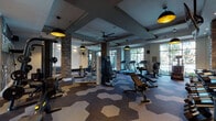 This fitness center delivers a polished, modern aesthetic supported by thoughtful architectural details throughout its spacious, single-level layout. The standout flooring features a bold hexagonal pattern in varying shades of gray and tan, lending a dynamic visual energy to the space. Structural brick-clad columns punctuate the floor plan, adding an industrial character that contrasts with the otherwise sleek, contemporary design.

The coffered ceiling incorporates recessed lighting alongside striking black-and-gold pendant fixtures, creating a layered lighting scheme that keeps the space well-illuminated without sacrificing ambiance. A ceiling fan provides additional airflow across the main workout area. Floor-to-ceiling windows along one wall flood the interior with natural light and offer views of the surrounding greenery, while a built-in seating bench with cubbies lines one wall for convenience.

The facility is comprehensively equipped with treadmills, cable machines, a multi-functional rack system, free weights with a full dumbbell rack, adjustable benches, and additional cardio machines. Full-length mirrors expand the sense of space and serve functional purposes throughout. A dedicated alcove features an artistic wall mural as a backdrop to a power rack, adding a creative focal point. A small refreshment station with a sink and water fountain completes the amenity.