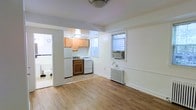 This single-floor space offers a functional and straightforward layout designed to maximize every square foot. The main living area features warm hardwood flooring that extends throughout, creating a cohesive and inviting atmosphere. White walls keep the space feeling bright and airy, complemented by natural light filtering in through multiple windows, one of which houses a window-mounted air conditioning unit for climate control. A cast-iron radiator provides supplemental heating, a classic feature that speaks to the building's character.

The kitchen is seamlessly integrated into the living area, separated by a partial wall that maintains an open feel. It is equipped with light wood cabinetry, a white gas range, a refrigerator, and a dark wood base cabinet that adds a subtle contrast to the overall palette. A ceiling-mounted light fixture illuminates the kitchen workspace. A doorway off the main area provides direct access to the bathroom, which is partially visible and includes a toilet, continuing the clean, utilitarian aesthetic of the space. Overall, this unit presents a practical and no-frills living environment with enduring architectural details.