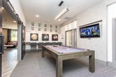 Located at Jennings Mill Parkway in Athens, Georgia, this single-floor game room is thoughtfully designed as a social and recreational amenity space. At its center sits a full-size pool table with a sleek, modern frame, surrounded by patterned carpet flooring that adds texture and warmth to the room. A large wall-mounted flat-screen TV is positioned prominently on a textured accent wall, making the space equally suited for watching sports or gaming. Seating is provided along one wall by a row of upholstered bar-height chairs set against a built-in counter, ideal for spectators or casual gatherings. The walls are decorated with bold billiard ball artwork and illuminated display niches showcasing decorative gold sculptural accents, lending the space a curated, gallery-like quality. A striking dark sliding barn door with a diagonal chevron pattern serves as both a functional partition and a bold architectural statement, connecting the game room to an adjacent area. Natural light filters in through large windows fitted with drapes, while recessed and track lighting overhead ensure the space is well-lit at any time of day. The overall design balances modern aesthetics with inviting, comfortable details, creating an engaging environment for recreation and socializing.