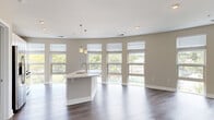 This contemporary single-floor residence showcases a thoughtfully designed open-concept layout that seamlessly connects the kitchen and living areas. A defining architectural feature is the sweeping curved exterior wall lined with floor-to-ceiling windows, flooding the space with natural light and offering expansive views of the surrounding urban streetscape and lush tree canopy.

The kitchen is well-appointed with white shaker cabinetry, light quartz countertops, a glass tile backsplash, and a full suite of stainless steel appliances including a gas range, built-in microwave, and French door refrigerator. A central island with an undermount sink and pendant lighting above serves as both a functional workspace and a natural gathering point. Rich dark hardwood-style flooring runs throughout the main living areas, adding warmth and contrast to the neutral palette.

The bedroom features plush wall-to-wall carpeting, large double-hung windows with views to the street below, and generous wall space. Two full bathrooms are finished with white cabinetry, quartz vanity tops, chrome fixtures, subway tile tub/shower surrounds, and wood-look luxury vinyl plank flooring. The overall aesthetic is clean, modern, and move-in ready, with a cohesive warm greige color scheme throughout.