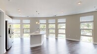 This contemporary single-floor residence showcases a thoughtfully designed open-concept layout that seamlessly connects the kitchen and living areas. A defining architectural feature is the sweeping curved exterior wall lined with floor-to-ceiling windows, flooding the space with natural light and offering expansive views of the surrounding urban streetscape and lush tree canopy.

The kitchen is well-appointed with white shaker cabinetry, light quartz countertops, a glass tile backsplash, and a full suite of stainless steel appliances including a gas range, built-in microwave, and French door refrigerator. A central island with an undermount sink and pendant lighting above serves as both a functional workspace and a natural gathering point. Rich dark hardwood-style flooring runs throughout the main living areas, adding warmth and contrast to the neutral palette.

The bedroom features plush wall-to-wall carpeting, large double-hung windows with views to the street below, and generous wall space. Two full bathrooms are finished with white cabinetry, quartz vanity tops, chrome fixtures, subway tile tub/shower surrounds, and wood-look luxury vinyl plank flooring. The overall aesthetic is clean, modern, and move-in ready, with a cohesive warm greige color scheme throughout.