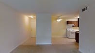This single-floor space offers a functional and straightforward layout that blends the living area and kitchen into one open, cohesive environment. The living area is finished with wall-to-wall carpeting in a warm neutral tone, creating a comfortable and understated foundation. A ceiling fan with a light fixture is centrally positioned, providing both ventilation and ambient lighting throughout the main space. A structural dividing half-wall partially separates the living zone, adding a subtle architectural element without fully closing off the open feel. The kitchen is tucked to one side and features dark wood cabinetry, a white electric range, and an overhead ventilation hood, offering a practical cooking space with efficient use of the available area. The entry door is visible from the main living area, contributing to the straightforward and accessible flow of the floor plan. Soft, warm lighting from flush-mount ceiling fixtures illuminates the space evenly. The walls are painted in a light yellow-beige tone, lending a neutral and welcoming ambiance throughout. The overall design emphasizes practicality and ease of living within a compact, well-organized single-level layout.