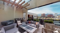 This elevated outdoor terrace showcases a thoughtfully designed rooftop living space that combines contemporary architecture with comfortable, functional outdoor amenities. The terrace is defined by crisp white brick exterior walls that frame the space and provide a clean, modern aesthetic backdrop throughout.

A prominent wooden pergola with exposed beam construction anchors the entertaining area, strung with globe lights that add warmth during evening gatherings. Beneath it, a centrally positioned glass-topped fire pit serves as the focal point of the seating arrangement, surrounded by an assortment of modern gray upholstered sofas, woven rope lounge chairs, and accent armchairs, all accented with bold orange throw pillows.

Large-format stone tiles cover the terrace floor, lending a refined, cohesive finish underfoot. Oversized metal planters filled with neatly trimmed boxwood shrubs provide natural greenery and subtle definition to the space. Expansive sliding glass doors connect the terrace directly to the interior, maximizing natural light and ensuring a seamless indoor-outdoor flow.

The terrace commands sweeping panoramic views of the surrounding cityscape and a distant mountain range, making it a standout feature of the space. The open-sky setting allows for abundant natural light throughout the day, enhancing the overall sense of openness and sophistication.