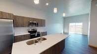 This contemporary single-floor unit showcases a thoughtfully designed open-concept layout with modern finishes throughout. The kitchen serves as the heart of the space, featuring a large center island with a white quartz countertop and double stainless steel sink, dark wood-tone cabinetry, and a full suite of stainless steel appliances including a side-by-side refrigerator, electric range, and over-the-range microwave. Pendant lighting hangs above the island, complementing the recessed ceiling lights and contributing to a bright, airy atmosphere. The kitchen flows seamlessly into the living area, where durable wood-look flooring and soaring ceilings create a spacious, welcoming feel, with access to an outdoor balcony providing natural light.

The bedrooms are carpeted for comfort and feature windows with exterior views, along with exposed metallic ductwork that lends an industrial character to the space. The primary bathroom is well-appointed with an elongated vanity, wood-tone cabinetry, white countertops, and wood-look tile flooring, with a separate water closet for added privacy. A walk-in closet with built-in shelving connects to the bedroom. A secondary half-bath offers a single vanity with a clean white countertop and mirror. A dedicated utility room houses the water heater and HVAC equipment, with a laundry hookup conveniently located nearby.