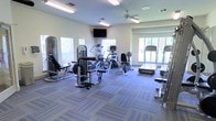 This fitness center offers a thoughtfully arranged workout space designed to accommodate a range of exercise needs. The room is bright and welcoming, with multiple large windows along the walls that allow generous natural light to fill the space while providing views of the surrounding greenery outdoors. A ceiling fan centered overhead aids in air circulation, contributing to a comfortable environment during workouts.

The flooring consists of durable, patterned carpet tiles in a blue-grey tone, providing cushioning underfoot throughout the space. Equipment is well-distributed across the room, including strength-training machines such as a cable/weight machine and a leg press, alongside a selection of cardio equipment including treadmills, an elliptical, a stationary bike, and a rowing machine. A wall-mounted flat-screen television is positioned for easy viewing from multiple points in the room, adding an entertainment element to workout sessions. A mirror on one wall enhances the sense of space and allows users to monitor their form. The overall ambiance is clean, functional, and inviting, making it a practical facility for daily fitness routines.