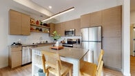 This contemporary single-floor residence showcases a thoughtfully designed open-concept layout with modern finishes and a warm, neutral palette throughout. The kitchen is a standout feature, boasting flat-front cabinetry in a natural wood tone, open floating shelves, a stone-topped island with built-in storage, stainless steel appliances, and a white tile backsplash. A linear pendant light anchors the space, adding a sleek architectural touch. Light hardwood flooring flows seamlessly through the main living areas, enhancing the sense of openness. The living room is bright and welcoming, featuring large picture windows that frame outdoor views and flood the space with natural light. The multiple bedrooms are well-proportioned, each with light hardwood floors and large windows. One bedroom includes a built-in sliding-door closet with wire shelving for practical storage. The bathrooms are finished with clean white countertops, undermount sinks, wood-framed mirrors, and glass-enclosed showers, blending functionality with modern elegance. The floating vanity in the primary bath adds a refined, contemporary detail. Overall, the residence offers a cohesive, modern aesthetic with quality finishes and a bright, airy ambiance throughout.