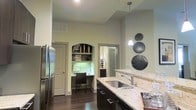 This single-floor residence showcases a well-appointed interior with a cohesive design aesthetic and attention to detail throughout. The kitchen serves as a central highlight, featuring dark espresso cabinetry, light granite countertops, a stainless steel sink, and a matching stainless steel refrigerator. A stylish pendant light hangs above the counter, adding a modern touch to the space. The walls are finished in a warm, neutral tone that complements the rich dark hardwood flooring, creating a grounded and inviting atmosphere. Soaring ceilings amplify the sense of openness and airiness throughout the main living area. A standout architectural feature is the built-in desk nook, framed by an elegant arched opening and equipped with open shelving above — an ideal dedicated workspace seamlessly integrated into the home. Decorative circular wall accents add a contemporary artistic element to the kitchen wall. Doorways leading to additional rooms suggest a well-organized floor plan with distinct private spaces. The overall ambiance is modern yet warm, balancing functional design with refined styling to create a comfortable and visually appealing living environment.