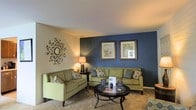 This single-floor residential space offers a thoughtfully designed interior that combines comfort and style. The living area serves as the heart of the home, featuring a bold navy blue accent wall that creates a striking focal point against the neutral, light-toned surrounding walls and ceiling. Two framed botanical art pieces are displayed prominently on the accent wall, adding a decorative touch that complements the overall color palette. A decorative starburst mirror adorns the adjacent wall, enhancing the room's visual interest. The living space is furnished with a pair of sage green sofas accented with patterned throw pillows, arranged around a classic round iron-and-glass coffee table, creating an inviting and functional seating arrangement. Warm ambient lighting is provided by a table lamp, contributing to the cozy atmosphere of the room. The floor is covered with neutral wall-to-wall carpeting that adds softness underfoot and ties the space together. The design aesthetic throughout is cohesive and welcoming, blending earthy greens, cool blues, and warm neutrals for a balanced and comfortable living environment suited for everyday living.