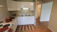 This single-floor space offers a clean and functional layout anchored by a well-appointed kitchen. The kitchen features classic white upper and lower cabinetry, providing ample storage space, paired with light-toned countertops that create a bright and open feel. A double-basin stainless steel sink sits centered beneath the upper cabinets, complemented by an under-cabinet light fixture that adds practical task lighting. The cabinetry's simple hardware and crisp white finish give the kitchen a timeless, understated aesthetic. Warm hardwood-style flooring extends throughout the kitchen and into the adjoining areas, lending a cohesive and inviting quality to the space. The soft, neutral green-gray wall color ties the kitchen together harmoniously. A colorful striped area rug anchors the kitchen workspace, adding a touch of warmth and character. A doorway visible from the kitchen leads to additional rooms, including what appears to be a hallway and closet space, suggesting a thoughtfully connected floor plan. Overall, the space conveys a practical and comfortable atmosphere suited for everyday living.