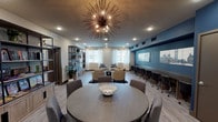 This thoughtfully designed single-floor community space seamlessly blends comfort, functionality, and modern aesthetics into one cohesive environment. The room is anchored by a striking sputnik-style chandelier that serves as a dramatic focal point, complementing the recessed lighting that illuminates the entire space with a warm, welcoming glow. Light wood-look flooring runs throughout, tying together the various distinct zones within the room.

A bold blue accent wall adds depth and visual interest, adorned with cityscape artwork that lends an urban sophistication to the space. Along this wall, a long built-in countertop workspace lined with rolling office chairs provides an efficient co-working station, ideal for productivity. Open metal and wood shelving units showcase curated books, plants, and decorative accessories, adding warmth and personality.

The social and lounge areas feature multiple seating arrangements, including a round dining table with upholstered chairs and a casual conversation grouping of neutral armchairs gathered around a low coffee table atop a vibrant area rug. Natural light filters in through windows dressed with sheer white curtains, enhancing the airy atmosphere. Together, these elements create a versatile, well-appointed space suited for both relaxation and collaboration.