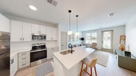 Located at 104 W Main St in Edmond, Oklahoma, this well-appointed 2-bedroom, 1-bathroom residence offers a thoughtfully designed single-floor layout that maximizes every square foot. The open-concept kitchen and living area serves as the heart of the home, anchored by a sleek white quartz kitchen island with a matte black faucet and bar seating. The kitchen features crisp white shaker cabinetry with black hardware, stainless steel appliances including a refrigerator, range, and built-in microwave, and light hardwood-style flooring. Two globe pendant lights hang above the island, adding a modern, airy touch. The living room benefits from large windows that invite generous natural light and outdoor views, creating a warm and inviting atmosphere. The primary bedroom offers a carpeted floor, built-in closet with shelving, and direct access to the hallway. The bathroom is finished with a white quartz vanity, shaker-style cabinetry with black hardware, and a large frameless mirror, maintaining the clean, contemporary aesthetic found throughout the residence. The overall design is cohesive and modern, with a neutral palette of whites and soft grays that lends the space a bright, airy ambiance.