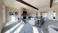 This thoughtfully designed single-floor clubhouse space offers an inviting and upscale atmosphere ideal for socializing and relaxation. The expansive main lounge serves as the centerpiece, featuring soaring ceilings accented by bold exposed dark wood beams that add rustic warmth to the otherwise contemporary design. A dramatic floor-to-ceiling stacked stone fireplace anchors the room and is topped with a wall-mounted television, creating a natural focal point. Plush seating arrangements, including sofas and chaise lounges, are arranged atop an intricately patterned area rug, fostering a comfortable and welcoming environment. A communal high-top table with bar-style seating provides an additional gathering area within the lounge. Large French doors along the perimeter allow generous natural light to pour into the space while offering views of the outdoors. Adjacent to the lounge is a well-appointed kitchen featuring rich dark wood cabinetry, granite countertops with a subway tile backsplash, stainless steel appliances, and an island with bar seating topped by an elegant chandelier. A dedicated café nook with a double sink and coffee station adds a charming and functional touch, making this space perfectly suited for hosted events and everyday community use.