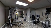 This single-level fitness room is designed to provide a functional workout environment within a relatively compact footprint. The space is finished with neutral taupe-grey walls and durable carpet tile flooring in a subtle pattern, contributing to a practical, no-frills aesthetic suited to an exercise setting.

Overhead, a drop ceiling with recessed panel lighting is complemented by a skylight that introduces natural light into the room, brightening the otherwise enclosed space. A decorative light fixture also hangs from the ceiling, adding a modest design accent.

The room is equipped with a multi-station cable weight machine positioned prominently on the left side, allowing for a variety of strength training exercises. Additional cardio equipment includes a treadmill, an upright stationary bike, and a recumbent bike, providing users with multiple options for cardiovascular workouts. Exercise balls are also present, adding versatility for core and balance training.

Large wall mirrors are strategically placed to visually expand the room and allow users to monitor their form during exercise. A small window on one wall provides a secondary source of natural light. A waste bin and wall-mounted accessories complete the practical setup of this well-equipped fitness space.