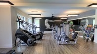 This single-floor fitness center is thoughtfully designed to support a comprehensive workout experience. The space is anchored by a striking black accent wall adorned with a large, dramatic mural of a figure in motion, creating an energizing and motivating atmosphere for users. Durable wood-look flooring runs throughout, offering both practicality and a clean, modern aesthetic.

The facility is outfitted with a variety of cardio equipment, including stair climbers and elliptical machines, positioned along one side of the room to maximize open floor space. A selection of Life Fitness strength training machines occupies the central area, providing options for targeted muscle training. Along the opposite wall, a full-length mirror expands the visual space and allows users to monitor their form during exercise. Additional fitness accessories, including stability balls and weighted balls, are visible in the reflected space, indicating a well-rounded selection of workout tools.

Natural light filters in through windows on one end of the room, complemented by recessed flat panel ceiling lights that ensure consistent, bright illumination throughout. A ceiling fan overhead contributes to air circulation and comfort during intense workouts. The overall ambiance is modern, clean, and purpose-driven, making this fitness center a functional and inviting space for daily exercise.