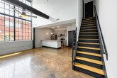 This two-floor unit at 11900 Main St in Fredericksburg, Virginia, blends industrial character with contemporary upgrades. The main level greets visitors with soaring ceilings, polished concrete floors, and a striking whitewashed exposed brick wall that anchors the open living area. Large factory-style windows flood the space with natural light, offering views of the surrounding brick architecture. A modern staircase with navy blue steel framing and wood treads leads to the upper level.

The kitchen is a standout feature, equipped with gray shaker cabinetry, granite countertops, a subway tile backsplash, and a full suite of stainless steel appliances including a refrigerator, range, microwave, and dishwasher. A granite-topped island with a deep sink and industrial-style faucet provides additional prep and dining space, illuminated by pendant lighting overhead.

The bathroom on the main level features a granite vanity with undermount sink and a large mirror, while a second full bathroom includes a soaking tub with floor-to-ceiling large-format tile surround. The upper level offers a bright private room with light wood laminate flooring, bold dark trim, and a window overlooking the interior of the building. This space masterfully combines raw industrial elements with polished, modern living essentials.