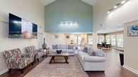 Located at 601 Jones Ferry Rd in Carrboro, NC, this single-floor community living space offers a welcoming and well-appointed environment designed for comfort and social interaction. The standout architectural feature is the dramatic vaulted ceiling that rises to impressive heights in the main seating area, accentuated by a bold teal accent wall and large clerestory windows that flood the space with natural light. Rich hardwood flooring runs throughout, lending warmth and continuity to the open layout. The main living area is generously arranged with multiple seating zones, including plush sofas, patterned accent chairs, and swivel lounge chairs centered around a wood coffee table atop a colorful area rug. A wall-mounted flat-screen TV and sleek track lighting add modern functionality. A secondary, lower-ceilinged zone features recessed lighting and additional flexible seating, providing a quieter, more intimate setting. A small kitchenette area is visible along one wall, offering convenient amenities. Multiple exterior doors—including a glass-panel entry and a rear door leading to an outdoor deck—ensure easy access and reinforce the bright, airy atmosphere. A restroom is accessible from within the space, enhancing its utility as a shared community hub.