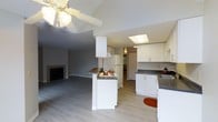 This single-floor residence offers a welcoming and functional layout centered around an open-concept living and kitchen area. The kitchen is well-appointed with white cabinetry, dark countertops, a built-in cooktop on a peninsula island, a white refrigerator, and a sink with ample counter space. A skylight overhead fills the kitchen with natural light, enhancing the bright, airy feel created by the crisp white finishes throughout. The peninsula serves double duty as a prep space and casual dining area, seamlessly connecting the kitchen to the adjoining living space. The living area is anchored by a classic tile-surround fireplace, adding warmth and a focal point to the room. A ceiling fan with a light fixture is centrally positioned, providing both comfort and illumination. Light wood-tone flooring extends throughout, lending a clean, cohesive look to the open layout. A doorway visible from the kitchen suggests additional rooms beyond, adding to the overall sense of a complete and practical floor plan. The neutral color palette of soft grays and whites throughout creates a calm, move-in-ready atmosphere suited to a variety of living styles.