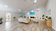 This single-floor community space exudes a relaxed, coastal aesthetic through its thoughtfully curated design and bright, airy atmosphere. Light wood-look flooring runs throughout, complementing the neutral gray-and-white color palette that gives the space a clean, welcoming feel.

The main lounge area serves as the centerpiece of the space, anchored by a shiplap accent wall featuring a built-in fireplace and a wall-mounted flat-screen TV above. Cream-toned sofas and accent chairs are arranged around a woven rug and natural wood coffee table, creating an inviting seating arrangement ideal for gathering. A ceiling fan with a pendant-style mount adds both function and style overhead, while recessed lighting ensures the space remains well-lit throughout.

Adjacent to the lounge is a more casual seating nook with two accent chairs, a small wood side table, and beach-inspired wall art rendered on reclaimed wood panels — a recurring decorative motif that reinforces the coastal theme. A dining area with a natural wood table and bench seating provides additional space for socializing.

Large double glass doors and expansive windows flood the interior with natural light and offer direct views of an outdoor pool area and landscaped grounds. The vaulted ceilings in this section of the space amplify the sense of openness. Lush potted plants are placed throughout, adding life and color to the overall design.