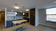 This single-floor space combines contemporary design with functional living. The kitchen is a standout feature, showcasing dark wood cabinetry that extends to the ceiling, providing generous storage. A mosaic tile backsplash adds texture and visual interest, while stainless steel appliances, including a range and microwave, offer modern convenience. A freestanding island with a butcher block and metal lower shelf serves as a versatile workspace and casual gathering spot. Pendant lighting and flush-mount ceiling fixtures illuminate the area warmly, and light wood-style flooring ties the space together cohesively.

A hallway connects the main living areas to the bathroom, which is finished with a granite-topped vanity set in gray cabinetry. The dual undermount sinks and large mirrors with overhead lighting create a bright, clean aesthetic. The neutral wall palette of warm whites and soft grays is consistent throughout, contributing to an open, airy atmosphere. Overall, the space reflects a modern design sensibility with quality finishes and thoughtful layout.