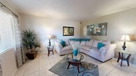 This efficiently designed single-floor residence offers a thoughtful layout encompassing a bedroom, two bathrooms, a combined kitchen and living area, a dedicated office, and a closet. The open-concept living room is finished with light-toned ceramic tile flooring and features a spacious sectional seating arrangement, a glass-top coffee table, and tasteful wall art that lends the space a relaxed, contemporary feel. Adjacent to the living area, the kitchen is equipped with dark wood cabinetry, granite countertops, a tile backsplash, and stainless steel appliances, complemented by a compact dining area with a round bistro-style table. The primary bedroom features soft carpeting, a ceiling fan, a sliding glass door that invites natural light, and a built-in closet for ample storage. A second carpeted room serves as a versatile office space with neutral walls and a ceiling light fixture. The two bathrooms are finished with large-format tile surrounds in the tub/shower areas and feature granite-topped vanities with dark wood cabinetry, adding a touch of refinement. Throughout the residence, a cohesive teal and neutral color palette creates a calm, welcoming ambiance.