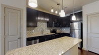 This single-story residence offers a well-designed layout with a strong emphasis on modern finishes and functional living spaces. The kitchen serves as a central highlight, showcasing rich dark cabinetry with sleek hardware, light granite countertops, and a mosaic tile backsplash in neutral earth tones. A large kitchen island with matching granite provides generous prep and dining space, illuminated by a series of pendant lights suspended from above. Stainless steel appliances, including a refrigerator, range, microwave, and dishwasher, complete the gourmet setup. Wood-style flooring runs through the kitchen and living areas, adding warmth and continuity. The open layout allows a clear sightline from the kitchen toward the living area, which benefits from natural light through a window and is equipped with a ceiling fan.

The bedrooms feature soft carpeting, ceiling fans, and windows with blinds, providing a comfortable and quiet retreat. The bathroom includes a soaking tub with tile surround, wood-style flooring, a dark vanity with granite countertop, and access to a walk-in closet with built-in shelving. Overall, the space presents a cohesive, contemporary aesthetic throughout.