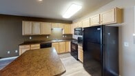 Located at 1720 13th St SE in Minot, North Dakota, this single-floor unit offers a practical and functional layout with well-defined living spaces throughout. The heart of the home is a well-appointed kitchen featuring light maple cabinetry, dark countertops, and a full suite of black appliances including a range, built-in microwave, dishwasher, and refrigerator. A central island with matching countertop provides additional prep space and casual seating. The kitchen opens directly into a generously sized living area finished with smooth hardwood-style flooring, recessed lighting, and a large sliding glass door fitted with vertical blinds that floods the space with natural light and provides access to the outdoors. The bedrooms are carpeted for comfort and warmth, with neutral-toned walls, baseboard heating, and windows that bring in natural light. The bathroom is clean and straightforward, featuring a full soaking tub with a shower, chrome fixtures, and wood-style flooring. Throughout the unit, warm earth-toned walls and consistent flooring choices create a cohesive and inviting atmosphere suited for comfortable everyday living.