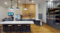 Located at 4400 SW 20th Ave in Gainesville, FL, this single-floor community amenity space is designed to foster social interaction and relaxation. The kitchen area is a standout feature, boasting dark gray cabinetry, white quartz countertops, and a large central island with bar seating. A shiplap wood accent wall serves as a striking backdrop, complemented by open shelving and industrial-style pendant lighting. Stainless steel appliances, including a full-size refrigerator, complete the functional yet stylish setup. The lounge area exudes a modern, eclectic character with a bold navy accent wall that rises to vaulted ceilings, a wall-mounted flat-screen TV, mid-century modern leather chairs, and a vibrant patterned area rug over warm hardwood-style flooring. Natural light filters in through large grid-pane windows, adding to the inviting ambiance. The game room extends the social atmosphere with a regulation pool table, an open shelving unit stocked with board games and plants, a ceiling-mounted projector, and a sunken lounge nook. Together, these spaces create a well-rounded, community-oriented environment ideal for gathering, entertainment, and everyday enjoyment.