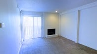 This single-story space centers around a bright, open living area designed with clean, understated finishes. The room's most notable feature is a white-painted brick fireplace positioned along the main wall, providing a classic focal point and a functional source of warmth. A raised brick hearth extends the fireplace's visual presence, lending the space a timeless, traditional character.

Natural light fills the room through a large sliding glass door fitted with full-length vertical blinds, which offer both privacy and light control. The door provides direct access to an outdoor area, enhancing the sense of openness within the space. A recessed ceiling light supplements the natural illumination, ensuring the room remains well-lit throughout the day.

The flooring throughout is covered in neutral-toned carpet, contributing to a soft, comfortable atmosphere. The textured ceiling adds subtle visual depth, while the smooth white walls and clean lines maintain a simple, cohesive aesthetic. A wide, open wall section adjacent to the fireplace offers flexible options for furniture arrangement or decorative use, making the living area both practical and adaptable.