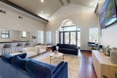 Located in Houston, TX, this community gathering space at 12430 Oxford Park Dr offers an inviting and well-appointed interior designed for comfort and socializing. The room immediately impresses with its dramatic vaulted ceilings and exposed dark wood beam accents, creating a sense of grandeur and openness. Rich hardwood flooring runs throughout, anchoring the space with warmth and character.

The main lounge area is furnished with plush navy blue velvet sofas and cream accent chairs arranged around a marble-top coffee table, all set atop a soft area rug. A wall-mounted flat-screen TV and a sleek credenza serve as a media focal point, while curated wall art and greenery add polished finishing touches. A bold navy accent wall adds a striking visual element.

Natural light pours in through multiple windows and a set of tall arched glass entry doors, offering outdoor views and an airy, welcoming atmosphere. Additional seating zones, including a bistro-style bar-height table with navy chairs and a casual dining nook, provide flexible arrangements for various activities.

A functional kitchenette area is tucked within the space, equipped with a double basin sink, dishwasher, microwave, and counter space, making the room well-suited for hosted events and gatherings. The overall ambiance is modern yet comfortable, blending upscale design with practical communal amenities.
