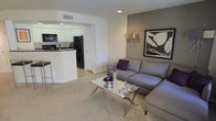This single-floor residence is designed with an open-concept layout that seamlessly connects the living area to the kitchen, creating a cohesive and functional flow. The living room is carpeted and centers around a sleek glass-topped coffee table with metallic legs, complemented by a contemporary gray sectional sofa accented with deep plum throw pillows. A large framed botanical artwork anchors one wall, paired with a modern arc floor lamp that adds warm ambient lighting. A recessed ceiling light provides additional illumination overhead.

The adjacent kitchen is visible through an arched passthrough opening and features white cabinetry, dark countertops, and black appliances including a refrigerator and microwave. A raised breakfast bar with seating separates the kitchen from the living space, making it ideal for casual dining or entertaining. Light tile flooring in the kitchen contrasts with the soft carpeting of the living area, subtly delineating the two zones. The overall color palette of warm beige, gray, and crisp white gives the space a clean, contemporary ambiance.